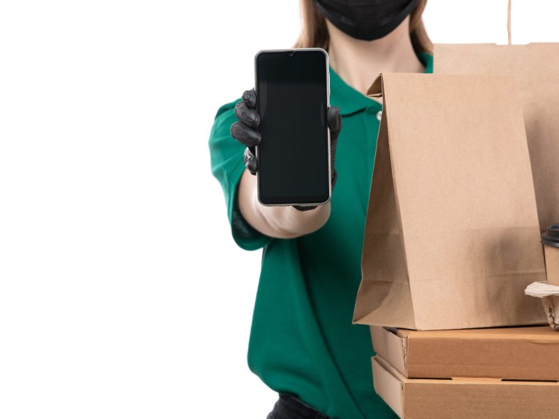 front-view-young-female-courier-green-uniform-black-gloves-black-mask-holding-food-delivery-packages-phone-delivering